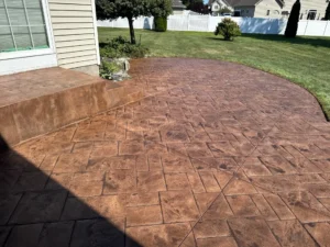 Recolored and resealed warm copper toned ashlar slate stamped concrete patio with matching step riser at a tan sided home with white vinyl fence, by NG Masonry serving Brighton NY