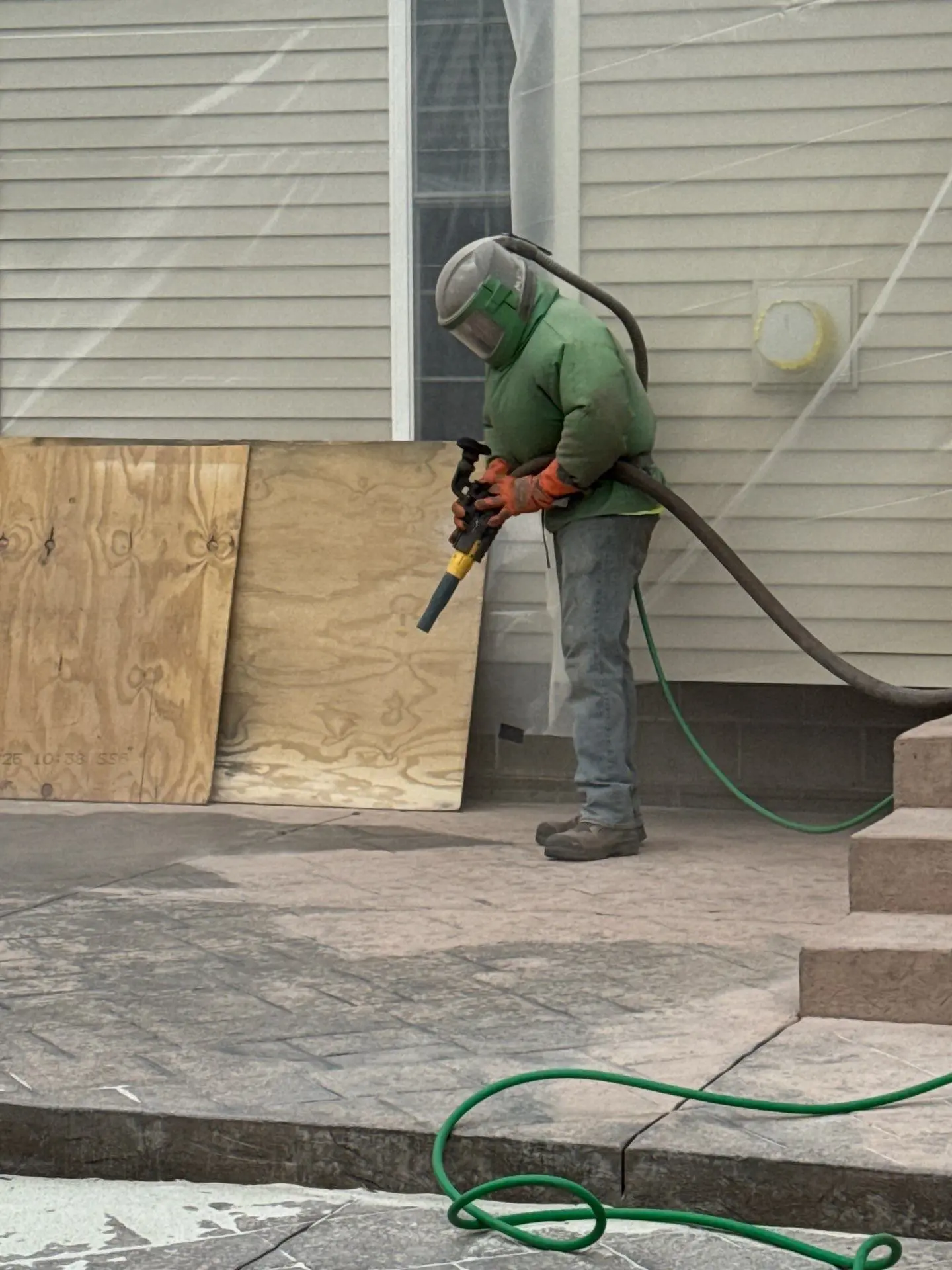 NG Masonry technician in face shield and respirator pressure spraying concrete prep on a covered porch slab beside a tan sided home, serving Greece NY