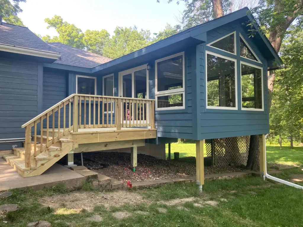 Newly built dark blue sided sunroom and elevated deck addition with pressure treated wood railing wrapping a ranch home in a wooded yard, by NG Masonry serving Rochester NY