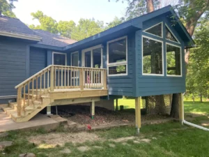 Newly built dark blue sided sunroom and elevated deck addition with pressure treated wood railing wrapping a ranch home in a wooded yard, by NG Masonry serving Rochester NY