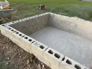 Rectangular concrete block foundation walls built on a smooth concrete slab in a grass field with wheelbarrow and generator on the perimeter, by NG Masonry serving Canandaigua NY