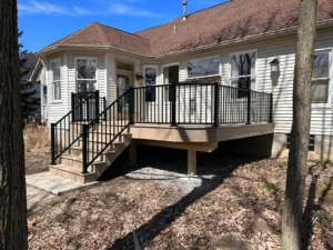 Tan composite deck with black aluminum railing and side stairs at the back of a white sided ranch home with bay window, by NG Masonry serving Batavia NY