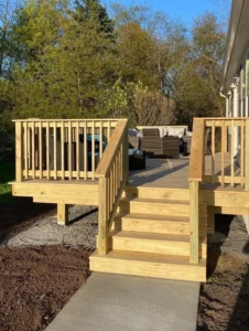 Newly built tan composite deck with pressure treated wood pickets and stairs leading down to a concrete walkway with wicker seating, by NG Masonry serving Chili NY