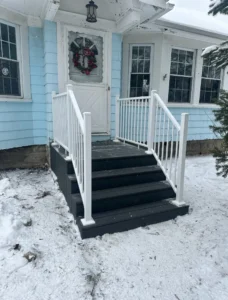 Winter view of a black composite deck with white aluminum railing and stairs at the front entry of a light blue sided home, by NG Masonry serving Fairport NY