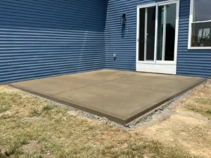 Sandy tan broom finish concrete patio slab at the back of a blue sided home, by NG Masonry serving Batavia NY