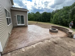 Tan stamped concrete patio with stone seat wall and round fire pit behind a tan sided home, by NG Masonry serving Batavia NY