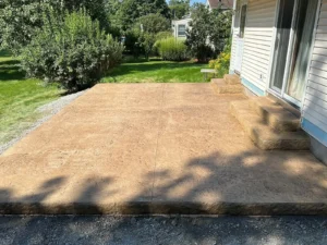Tan broom finish concrete patio with stamped border at the back of a cream sided home, by NG Masonry serving Brighton NY