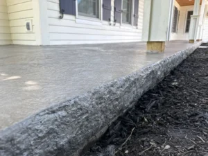 Closeup of a light gray broom finish concrete patio with rough rock faced edge beside a white sided home, by NG Masonry serving Brighton NY