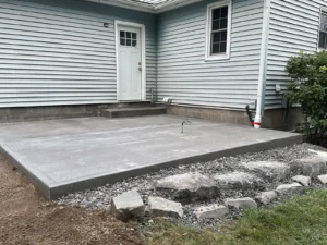 Square gray broom finish concrete patio with door step at a light blue ranch home, by NG Masonry serving Brockport NY