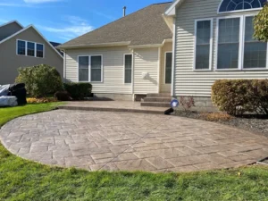 Round tan ashlar slate stamped concrete patio with charcoal border at a cream sided ranch home, by NG Masonry serving Chili NY