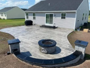 Wide light gray broom finish concrete patio with charcoal stamped border and round stone fire pit at a ranch home, by NG Masonry serving Fairport NY