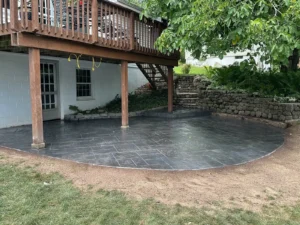 Charcoal slate textured stamped concrete patio under a wood deck with stone retaining wall, by NG Masonry serving Gates NY