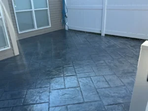 Charcoal slate textured stamped concrete patio in a herringbone pattern between siding and a vinyl fence, by NG Masonry serving Geneva NY