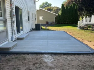Light gray broom finish concrete patio with charcoal stamped border at a white sided home, by NG Masonry serving Henrietta NY
