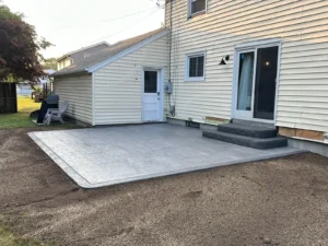 Square light gray broom finish concrete patio with two step risers at a cream sided home, by NG Masonry serving Irondequoit NY