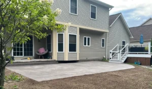 Light gray broom finish concrete patio at a gray sided two story home with white deck staircase, by NG Masonry serving Mendon NY
