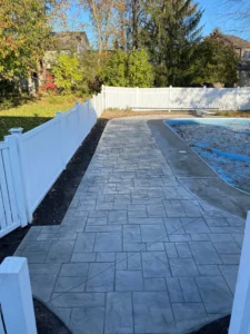 Light gray ashlar slate stamped concrete walkway alongside a pool with white vinyl privacy fence, by NG Masonry serving Newark NY