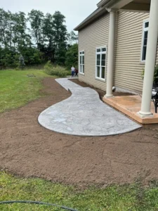 Curving large medallion stamped concrete walkway in a light gray flagstone pattern leading from a tan sided home porch over freshly graded soil, by NG Masonry serving Canandaigua NY