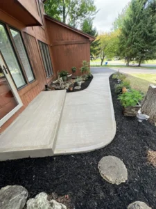 Wide light tan broom finished concrete walkway with a curved end wrapping around a dark stained wood sided home with landscape plantings, by NG Masonry serving Chili NY