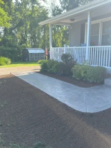 Light gray broom finished concrete sidewalk wrapping around the covered front porch of a white sided home with an American flag, by NG Masonry serving East Rochester NY