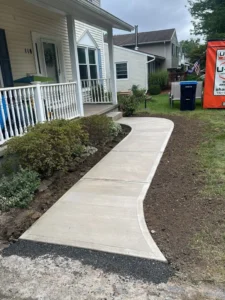 Curving light gray broom finished concrete walkway leading from a white wood porch out to the street with freshly graded soil on either side, by NG Masonry serving Fairport NY