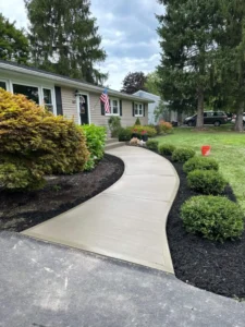Curving light tan broom finished concrete walkway through landscaping leading to the front entry of a tan sided ranch home with an American flag, by NG Masonry serving Farmington NY