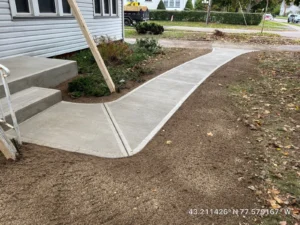 Wide light gray broom finished concrete walkway with step risers leading from a white sided home to the street through a freshly graded yard, by NG Masonry serving Geneva NY