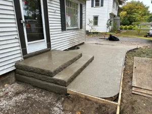 Wide broom finished concrete walkway with rough rock faced step risers leading from the side entry of a white sided home, by NG Masonry serving Greece NY