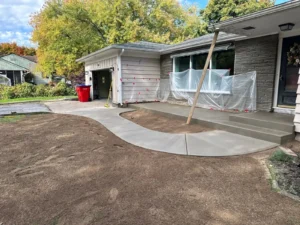 Curving light gray broom finished concrete walkway from a stone veneer ranch home entry around to the driveway, by NG Masonry serving Henrietta NY