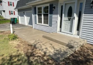 Light gray broom finished concrete walkway with step riser at the front entry of a gray sided two car garage home, by NG Masonry serving Hilton NY