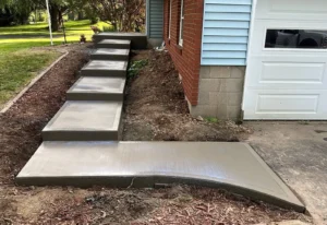 Stepped concrete walkway with multiple rise platforms transitioning a slope to a side entry beside a red brick and blue sided home, by NG Masonry serving Mendon NY
