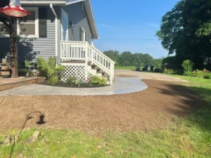 Curving light gray broom finished concrete walkway from a porch lattice down to the lawn beside a gray sided home, by NG Masonry serving Newark NY