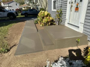 Wide L shaped broom finished concrete walkway with matching front step risers leading from the driveway to the entry of a gray sided home, by NG Masonry serving Penfield NY
