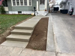 Wide light gray broom finished concrete walkway with matching front step risers leading up from the driveway to a green sided home with an American flag, by NG Masonry serving Rochester NY