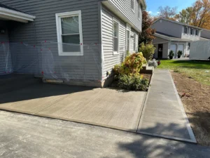 Wide light gray broom finished concrete walkway and pad alongside a gray sided two story home with adjoining sidewalk panels, by NG Masonry serving Victor NY
