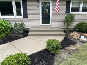 Light tan broom finished concrete front entry stoop with two step risers and curved end leading from the walkway to a tan sided ranch home, by NG Masonry serving Webster NY