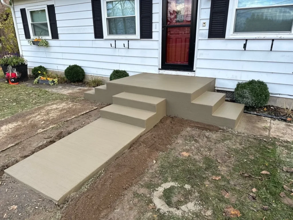 Tan broom finished concrete walkway with multi tier step risers leading up to a freshly poured stoop landing at a white sided home with red front door, by NG Masonry serving Williamson NY