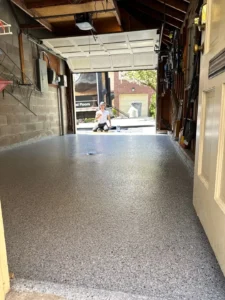 View down a single car garage with concrete block walls and a finished gray flake epoxy floor coating with a worker kneeling at the open garage door, by NG Masonry serving Canandaigua NY