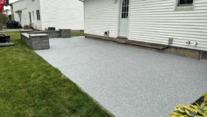 Wide outdoor concrete patio with gray flake polyaspartic floor coating wrapping behind a white sided home with stone seat wall pillars, by NG Masonry serving Chili NY