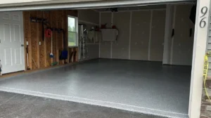 Two car garage with door open showing a finished gray flake epoxy floor coating, OSB wall on the left with tools hanging and drywalled back wall, by NG Masonry serving East Rochester NY