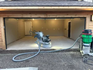 Concrete grinder set up at the threshold of a two car garage with cedar trim and dust vacuum hose, prepping the floor for epoxy coating, by NG Masonry serving Geneva NY
