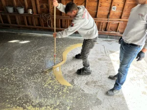 NG Masonry installer pouring tan tinted epoxy base coat onto a prepped concrete floor inside an old wood barn workshop, serving Greece NY