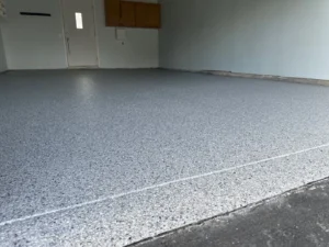 Finished gray flake epoxy garage floor with bright topcoat reflecting overhead light, side entry door and wood cabinets at back, by NG Masonry serving Irondequoit NY