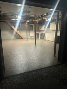 Night view into a basement with finished gray flake epoxy floor coating, steel support posts, wood stair landing, and white cabinetry, by NG Masonry serving Mendon NY