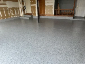 Finished gray flake epoxy garage floor coating in a renovated garage with unfinished drywall walls, side door, and stair landing, by NG Masonry serving Rochester NY