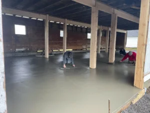 NG Masonry crew hand troweling a wide freshly poured concrete slab inside an open bay structure with wood post framing, serving Brighton NY