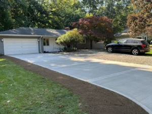 Wide light gray concrete driveway with darker apron leading to a white two car garage at a tan ranch home, by NG Masonry serving Canandaigua NY