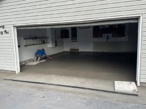 NG Masonry worker hand troweling a freshly poured smooth concrete floor inside a two car garage with linear drain at the threshold, serving Henrietta NY
