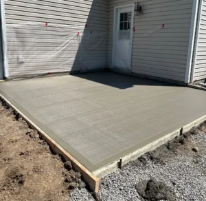 Freshly poured broom finished concrete shed pad with wood form boards still in place next to the back door of a white sided home, by NG Masonry serving Fairport NY