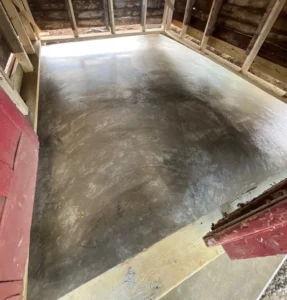 Wide freshly poured smooth gray concrete shed floor pad viewed from the door opening of a wood framed barn shed under construction, by NG Masonry serving Farmington NY
